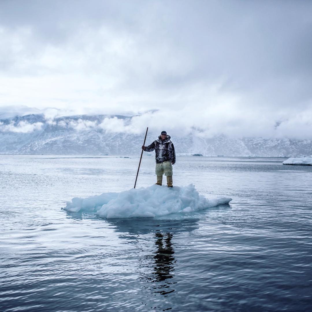 A man wearing a thick coat and pants stand on a floating ice berg in a lake surrounded by large icebergs
