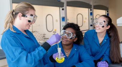 Three women wearing blue lab coats, goggles, and purple gloves are in the lab conducting experiments.