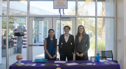 Three women stand behind a table with a sign that reads "Whittier College Established 1887" 