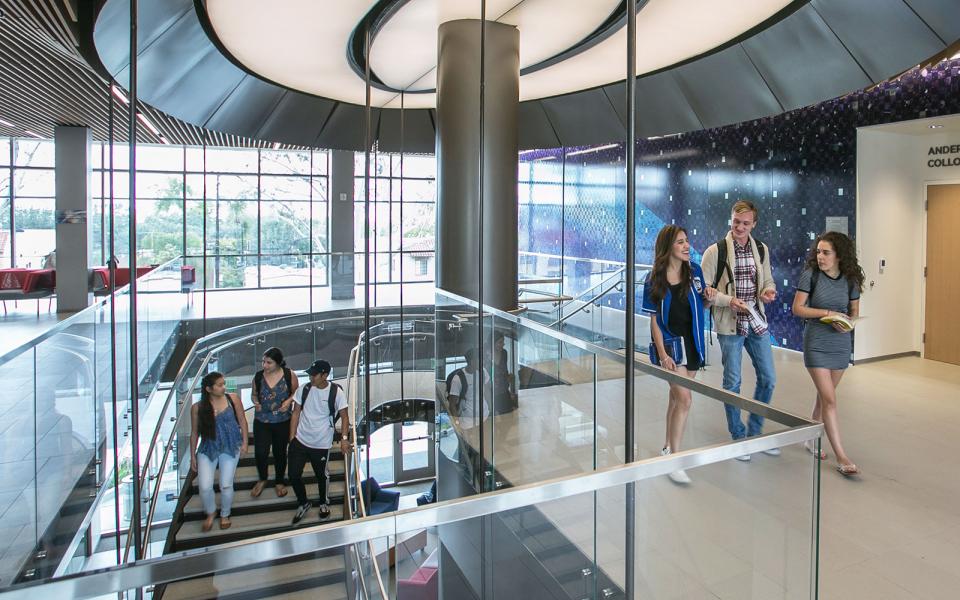 Three students walk down the stairs from the second floor to the first floor of the Science and Learning Center, while three students walk out of the second-floor lobby.